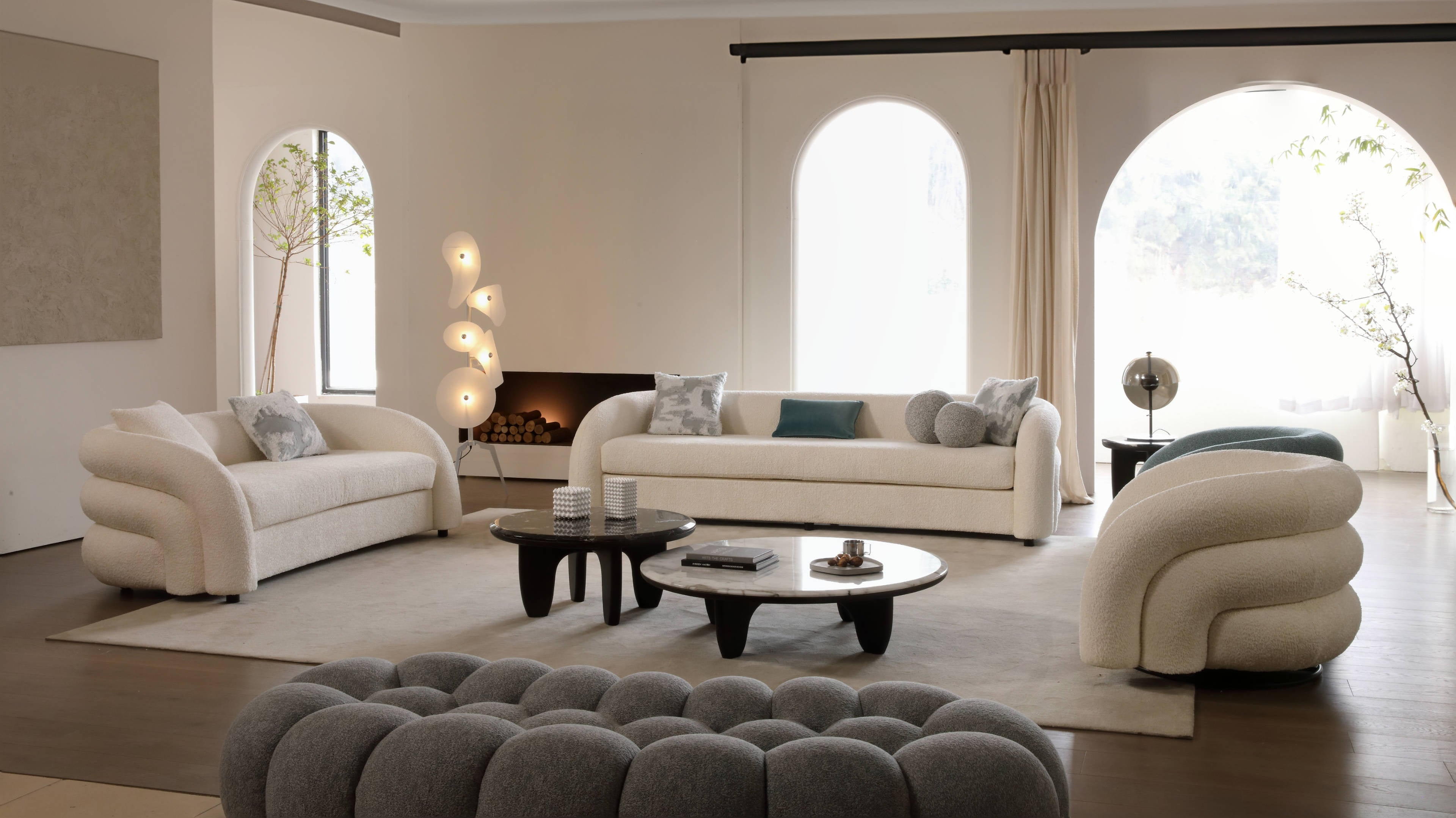 A cozy modern living room with plush sculptural sofas, a round coffee table, unique decorative lamps, and large arched windows.