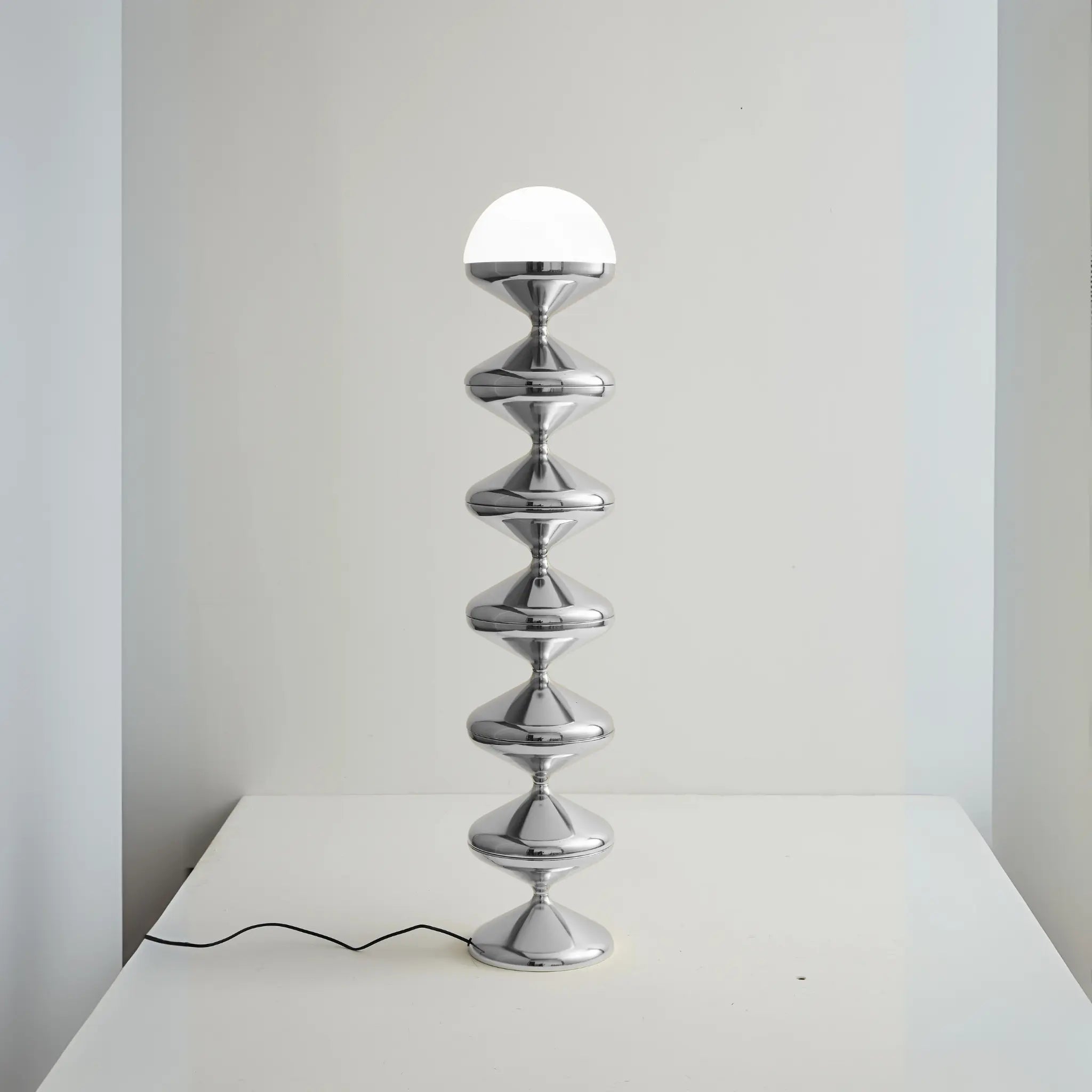 Modern chrome table lamp with stacked hourglass-shaped segments and a glowing white half-sphere bulb, placed on a white surface.