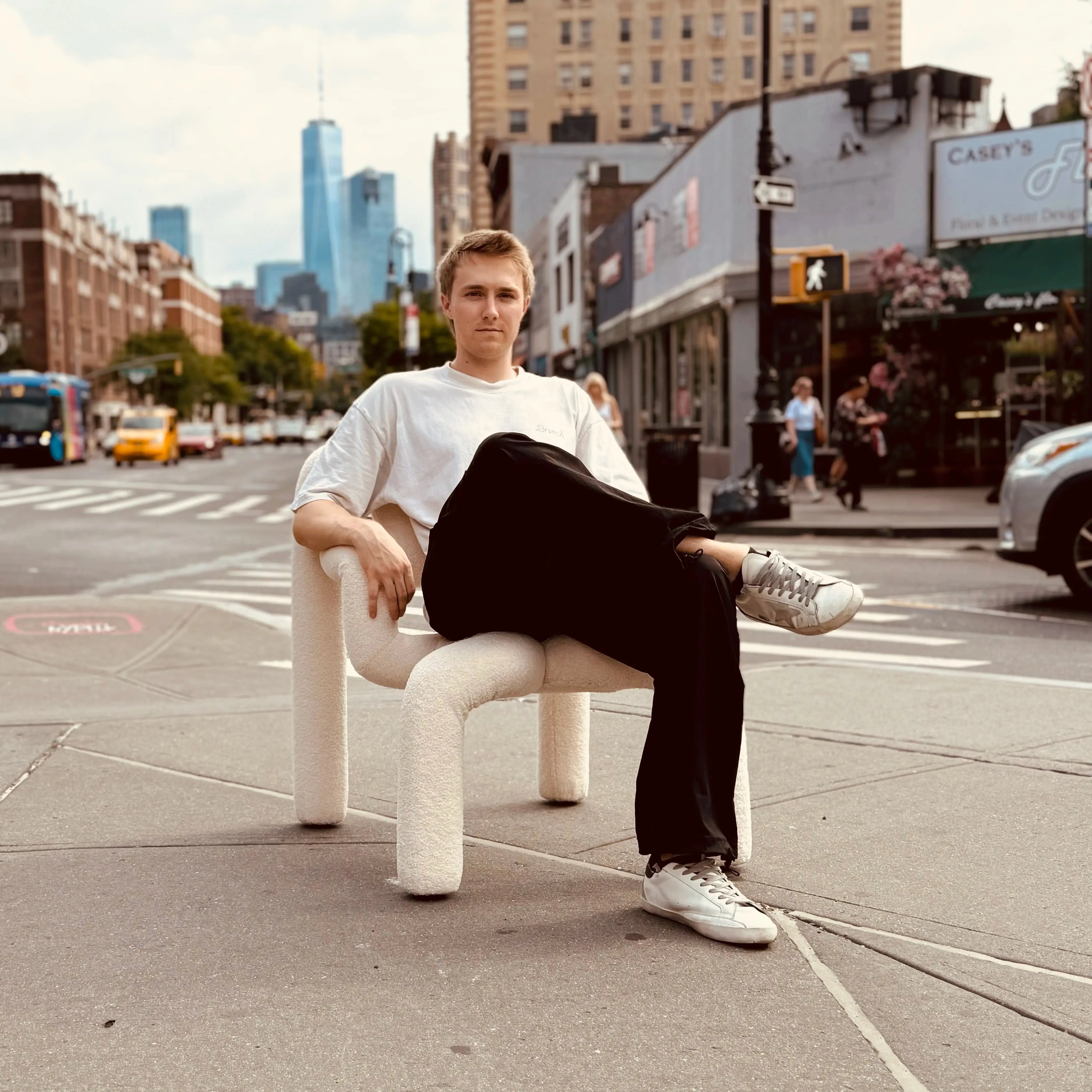 A person sits casually on a unique, plush chair in an urban setting, with city buildings and vehicles visible in the background.