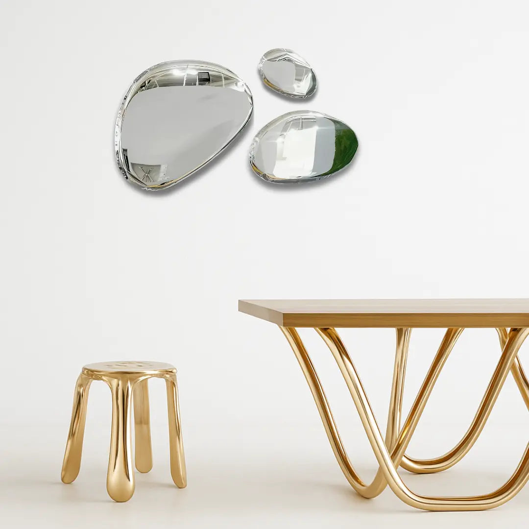 Modern interior with sculptural gold stool and wavy-legged gold table, set against a white wall with three organic-shaped silver mirrors.